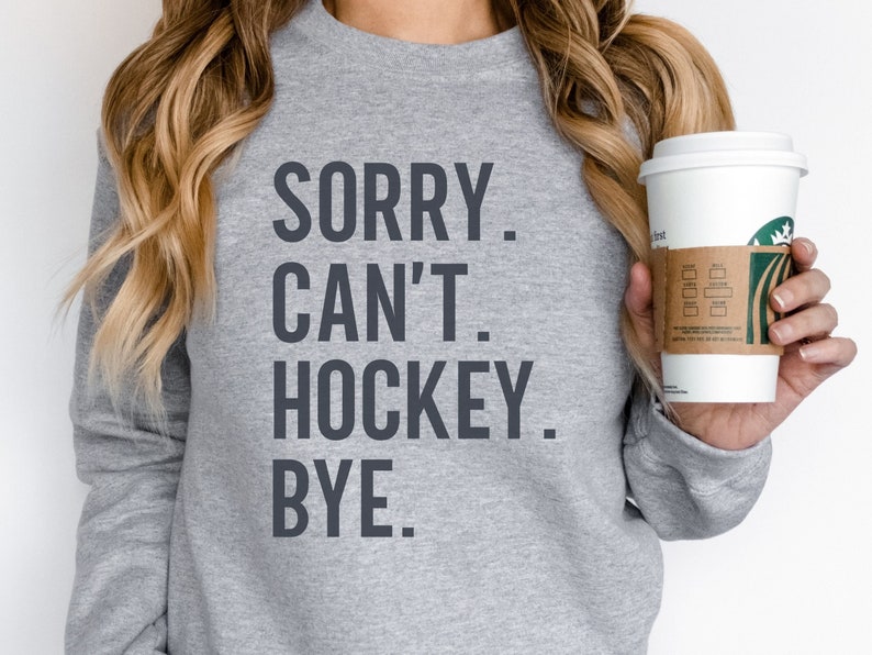 May include: Gray sweatshirt with the text "SORRY. CAN'T. HOCKEY. BYE." printed on the front. A person is holding a white coffee cup with a green logo.