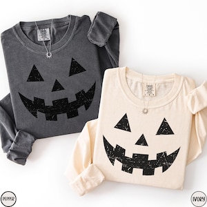 May include: Two long-sleeved shirts, one pepper gray and one ivory, each featuring a distressed black jack-o'-lantern face graphic. The shirts have a crew neck and are made of a soft material. A silver necklace is visible on each shirt.