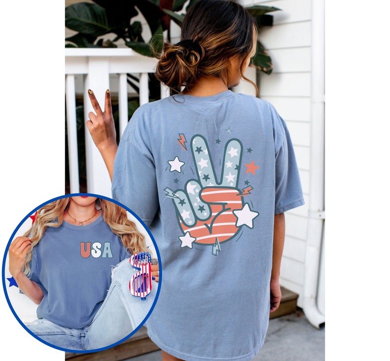 May include: A light blue t-shirt with a peace sign graphic on the back. The peace sign is red, white, and blue and has stars on it. The front of the shirt has the word "USA" in red, white, and blue letters.