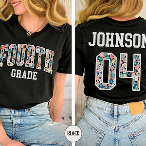 May include: Black t-shirt with "FOURTH GRADE" in floral print lettering on the front. The back of the shirt displays "JOHNSON 04" with floral print accents. The shirt is paired with blue jeans.
