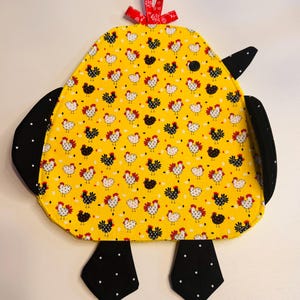 May include: A yellow chicken-shaped potholder with black and white polka dot wings and feet. The body is patterned with black, white, and red chickens on a yellow background. A red ribbon is attached at the top.