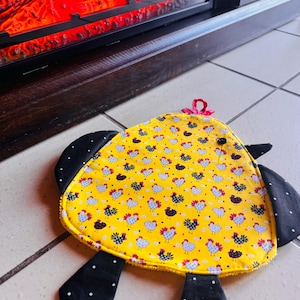 May include: A yellow, chicken-shaped pot holder with black polka-dot wings and feet. The pot holder is decorated with a repeating pattern of chickens and floral designs. A small red bow is attached to the top.