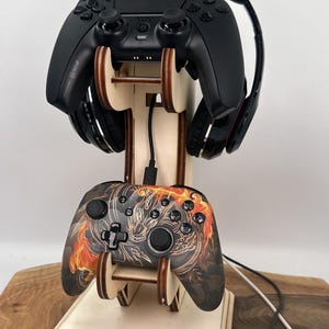 Puede incluir: Un soporte de madera con dos mandos de videojuegos y unos auriculares. El soporte está hecho de madera clara y tiene un acabado natural. Los mandos son negros y tienen una superficie texturizada. Los auriculares son negros y tienen detalles rojos. El soporte está diseñado para mantener los mandos y los auriculares en posición vertical.