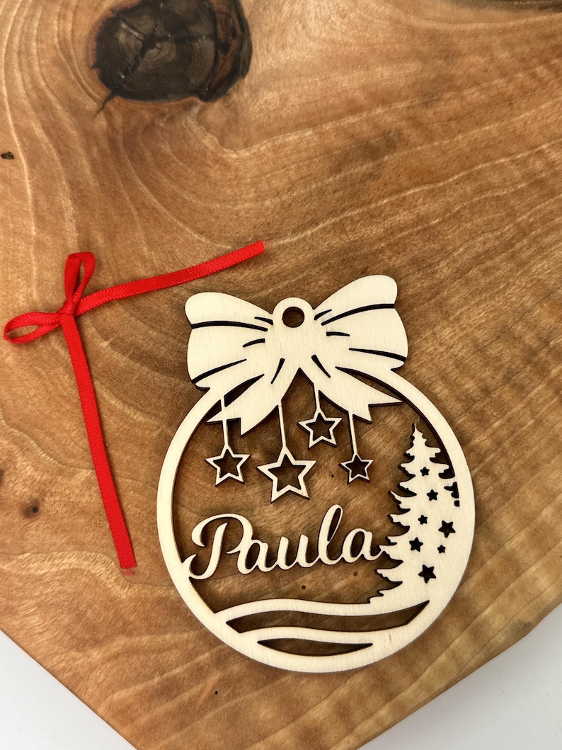 May include: A wooden Christmas ornament shaped like a bauble with a bow on top. The ornament has a tree and stars inside and the name "Paula" written on it.
