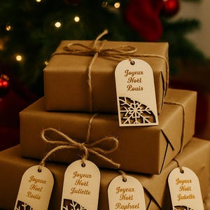 Könnte beinhalten: Gestapelte, braun verpackte Geschenke, mit Schnur gebunden. Jedes Paket hat ein Holzschild mit den Worten "Joyeux Noël" und einem Namen sowie einem Schneeflocken-Design. Die Geschenke sind vor einem Weihnachtsbaum angeordnet.
