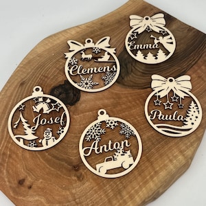 Könnte beinhalten: Fünf hölzerne Weihnachtskugeln mit verschiedenen Designs und Namen. Die Kugeln sind kugelförmig und haben oben eine Schleife. Die Namen auf den Kugeln sind "Clemens", "Emma", "Josef", "Anton" und "Paula".