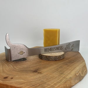Könnte beinhalten: Ein Bienenwachsschaber aus Edelstahl mit einem gebogenen Griff und einem kleinen Loch. Der Schaber trägt den Text "BEE4BUSINESS" und ein eingraviertes Bienendesign. Der Schaber liegt auf einem kleinen Holzscheit mit einem Block Bienenwachs darauf.