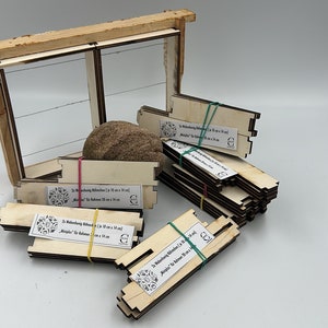 Può includere: Componenti in legno per telai apistici, inclusi telai assemblati e pile di pezzi non assemblati. I pezzi sono tenuti insieme da elastici. Il testo sulle etichette recita "2x Wabenhonig Rähmchen (je 10 cm x 14 cm) Miniplus für Rahmen 20 cm x 14 cm".
