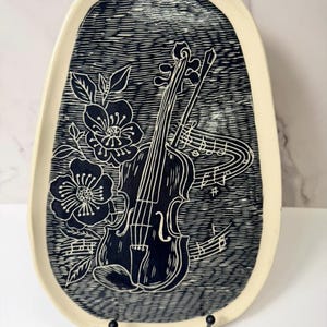 May include: A decorative, oval-shaped serving platter with a cream-colored border. The platter features a dark blue and white design of a violin, musical notes, and floral elements. The platter is displayed on a black metal stand.