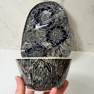 May include: A ceramic bowl and a matching upright plate featuring a dark navy blue glaze with carved white floral patterns. The bowl has a textured, vertical line design, while the plate displays three large, detailed flowers with leaves.