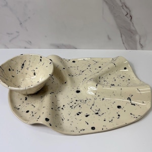 May include: Cream-colored ceramic serving dish and bowl set with black splatter design. The wavy-edged platter has a built-in dip bowl. A unique, handmade piece for serving appetizers or snacks.