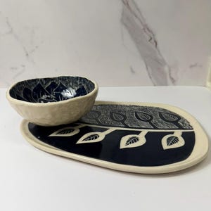 May include: A ceramic serving set featuring a small bowl and an oval platter. The bowl and platter have a cream-colored exterior and a navy blue interior. The navy blue interior of the bowl has a floral design. The platter has a leaf design.