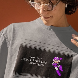 May include: A gray t-shirt with a pixelated graphic of a video game character and the text "You got debilitating anxiety".