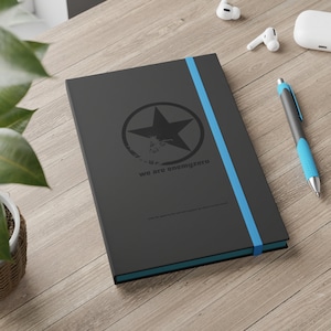 May include: A black notebook with a blue elastic band closure. The cover features a distressed black star inside a circle with the text "we are enemyzero".