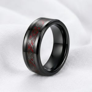 May include: A black ring featuring a polished finish and a wide center band with an intricate, raised black dragon pattern set against a vibrant red background. The band has beveled edges and a smooth, rounded interior surface for a comfortable fit.