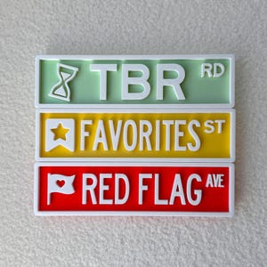 May include: Three colorful street sign plaques: TBR RD in green, FAVORITES ST in yellow, and RED FLAG AVE in red. Each sign has white lettering and a white border. The TBR sign features an hourglass icon.