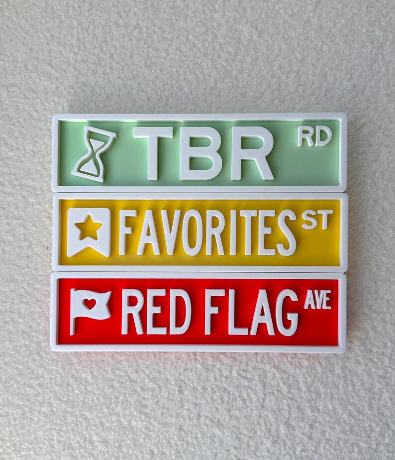 May include: Three colorful street sign plaques: TBR RD in green, FAVORITES ST in yellow, and RED FLAG AVE in red. Each sign has white lettering and a white border. The TBR sign features an hourglass icon.