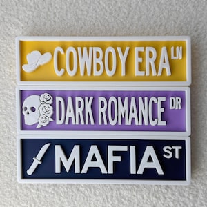 May include: Three decorative street signs with white lettering and borders. The top sign is yellow and reads "COWBOY ERA LN" with a cowboy hat graphic. The middle sign is purple and reads "DARK ROMANCE DR" with a skull and rose graphic. The bottom sign is navy and reads "MAFIA ST" with a knife graphic.
