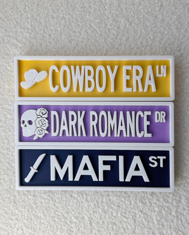 May include: Three decorative street signs with white lettering and borders. The top sign is yellow and reads "COWBOY ERA LN" with a cowboy hat graphic. The middle sign is purple and reads "DARK ROMANCE DR" with a skull and rose graphic. The bottom sign is navy and reads "MAFIA ST" with a knife graphic.
