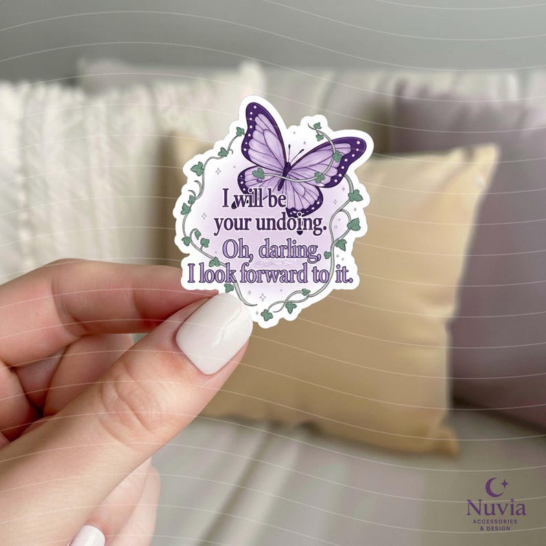 May include: A sticker featuring a purple butterfly with dark purple accents and green vines. The sticker has the text "I will be your undoing. Oh, darling, I look forward to it." The sticker is held by a person.