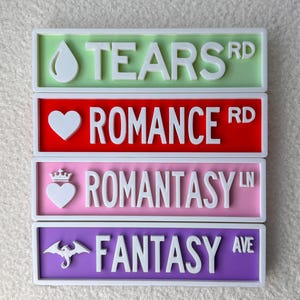 May include: Four colorful street signs with white lettering. The top sign is green and reads "TEARS RD" with a teardrop icon. The second sign is red and reads "ROMANCE RD" with a heart icon. The third sign is pink and reads "ROMANTASY LN" with a heart and crown icon. The bottom sign is purple and reads "FANTASY AVE" with a dragon icon.