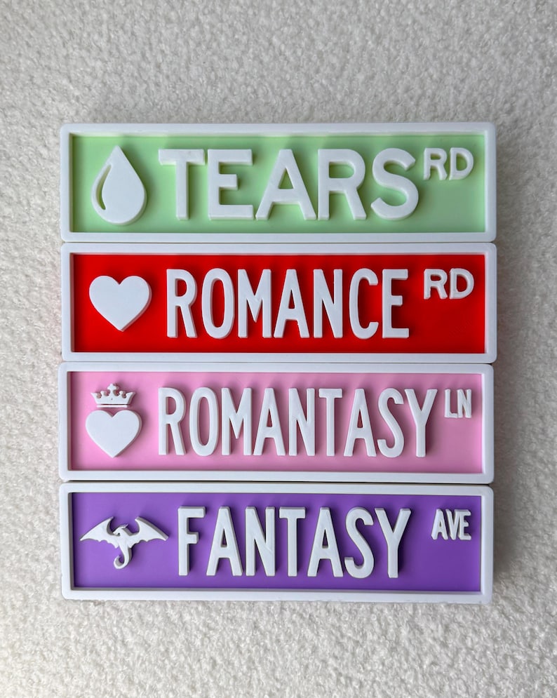 May include: Four colorful street signs with white lettering. The top sign is green and reads "TEARS RD" with a teardrop icon. The second sign is red and reads "ROMANCE RD" with a heart icon. The third sign is pink and reads "ROMANTASY LN" with a heart and crown icon. The bottom sign is purple and reads "FANTASY AVE" with a dragon icon.