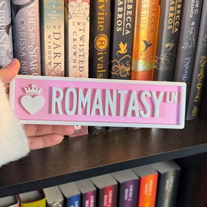 May include: A pink sign with the word "ROMANTASY LN" in white, featuring a heart and crown design. The sign is held in front of a bookshelf filled with books, including titles like "Divine Rivals" and "The Serpent & The Star".