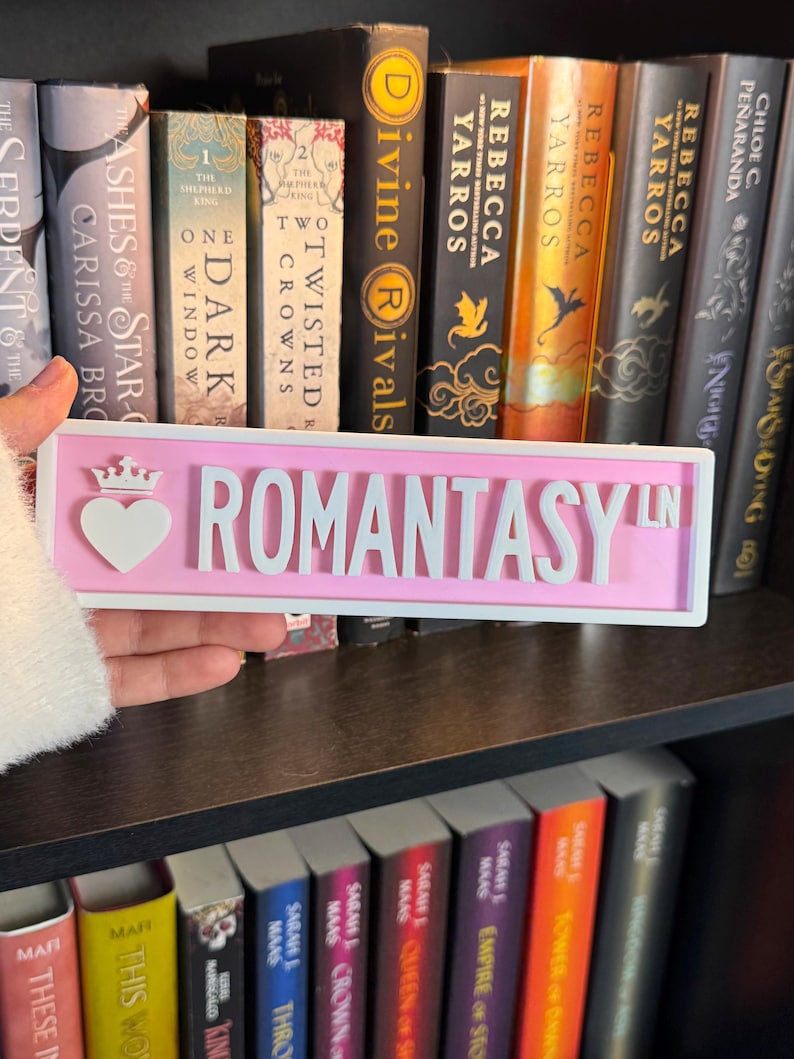 May include: A pink sign with the word "ROMANTASY LN" in white, featuring a heart and crown design. The sign is held in front of a bookshelf filled with books, including titles like "Divine Rivals" and "The Serpent & The Star".