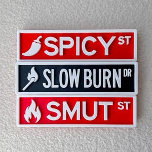 May include: Three novelty street sign magnets. The top sign is red with the text "SPICY ST" and a white chili pepper. The middle sign is black with "SLOW BURN DR" and a white match. The bottom sign is red with "SMUT ST" and a white flame.