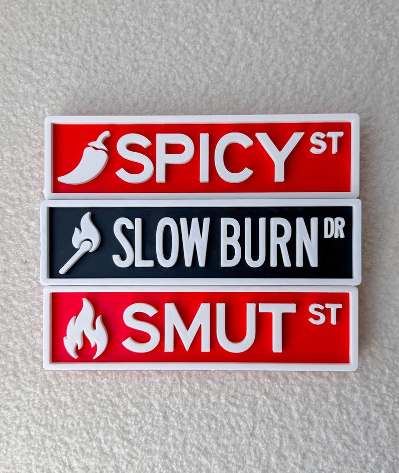 May include: Three novelty street sign magnets. The top sign is red with the text "SPICY ST" and a white chili pepper. The middle sign is black with "SLOW BURN DR" and a white match. The bottom sign is red with "SMUT ST" and a white flame.