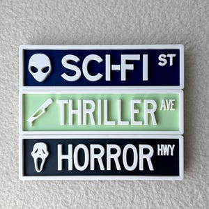 May include: Three rectangular signs with white borders. The top sign is navy blue with a white alien head and the text "SCI-FI ST". The middle sign is light green with a white knife and the text "THRILLER AVE". The bottom sign is black with a white ghost face and the text "HORROR HWY".