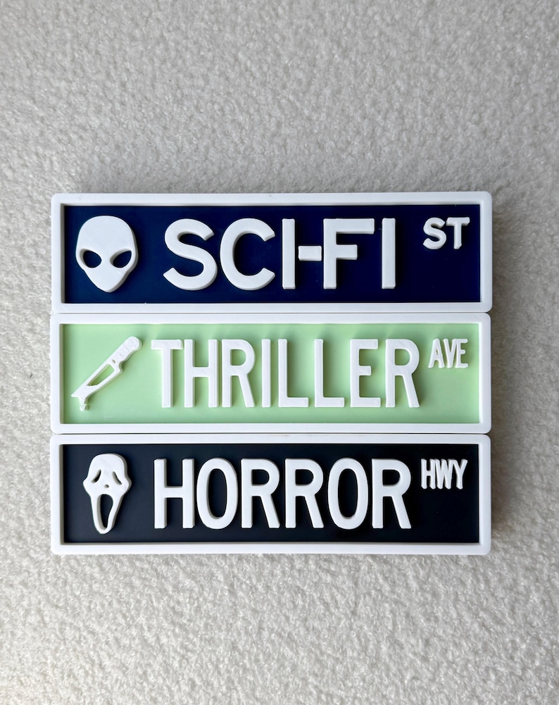 May include: Three rectangular signs with white borders. The top sign is navy blue with a white alien head and the text "SCI-FI ST". The middle sign is light green with a white knife and the text "THRILLER AVE". The bottom sign is black with a white ghost face and the text "HORROR HWY".