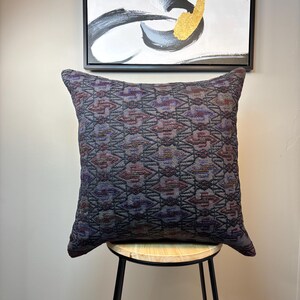 May include: A decorative pillow with a geometric pattern in shades of purple, gray, and brown. The pillow is sitting on a wooden stool with black metal legs.