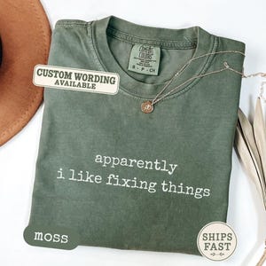 May include: A moss green short-sleeved t-shirt with the white text "apparently i like fixing things" printed on the front. The shirt features a crew neckline and a Comfort Colors brand label at the inner collar. A thin gold necklace rests on the fabric.