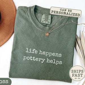 May include: A moss green crew neck t-shirt featuring the white text "life happens pottery helps" printed on the chest. The shirt has a Comfort Colors brand label at the inner neckline and is displayed with a gold necklace and a brown hat.