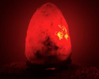 Op de afbeelding: Een grote, roze Himalaya-zoutlamp gloeit met een warm, oranje licht. De lamp heeft de vorm van een afgeronde, onregelmatige steen en staat op een donker, getextureerd oppervlak.