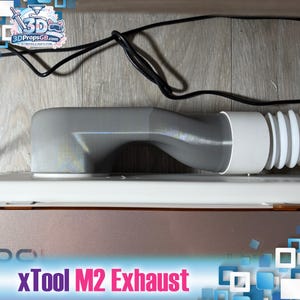 Könnte beinhalten: Ein grauer, 3D-gedruckter Abluftadapter für eine xTool M2 Lasermaschine mit einem gebogenen, starren Kunststoffkanal, der mit einem weißen flexiblen Schlauch verbunden ist. Die Einheit liegt auf einem metallisch braunen Gehäuse mit schwarzem Kabel.