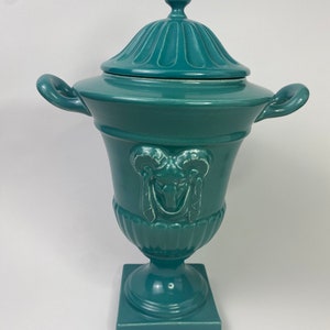 May include: Teal ceramic urn with a lid and a ram's head design. The urn has a square base and two handles.