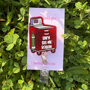 May include: A red and glittery badge reel featuring a medical cart design with the text "THEY SEE ME ROLLIN". The badge reel has a clear plastic clip and a retractable cord. The background is a white card with the shop's name.
