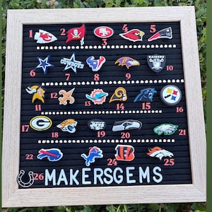May include: A framed black felt board displaying 26 colorful sports team logo charms. The logos include teams like the Cowboys, Raiders, and Chiefs. The text "MAKERSGEMS" is at the bottom. The frame is light-colored.