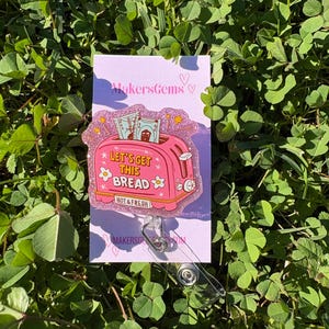 May include: A pink and glittery badge reel featuring a toaster with the text "LET'S GET THIS BREAD" and money popping out. The badge reel is on a purple card with the text "MakersGems".