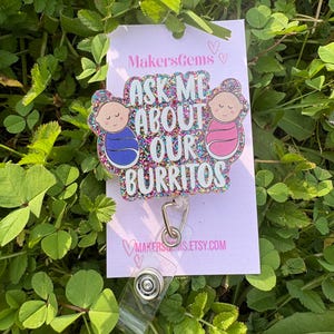 May include: A badge reel with the text "Ask Me About Our Burritos" and two baby illustrations. The badge reel is glittery and is on a pink card with the MakersGems logo and a clear plastic strap.