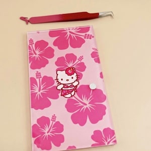 May include: A pink and red eyelash extension tweezer rests above a pink case with a floral pattern and a Hello Kitty graphic. The case has a white button closure. The floral pattern is a darker shade of pink.