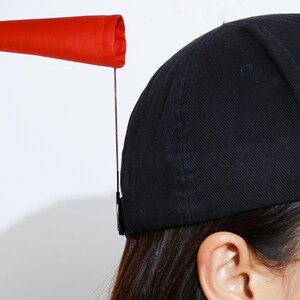 May include: A black baseball cap with a red windsock attached. The windsock is a bright red color and is attached to the cap with a thin metal rod. The cap is made of a dark fabric.