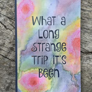 May include: A notebook with a watercolor design in shades of pink, yellow, blue, and purple. The cover features the text "What a Long Strange Trip It's Been" in black lettering. The notebook has a dark blue spine.