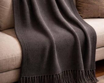 May include: A dark gray knitted throw blanket with fringe detail draped over a beige sofa. The blanket has a textured weave and is folded to show its full length. A brown pillow is visible on the sofa.