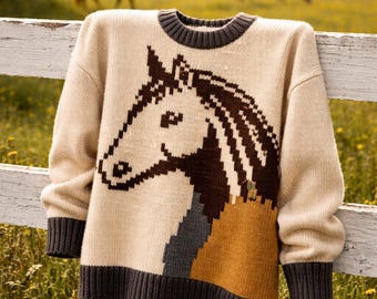 May include: A beige sweater with a horse head design. The horse's head is brown and white, with a brown mane and a tan and gray body. The sweater has a dark gray collar, cuffs, and hem. The sweater is hanging on a white fence.