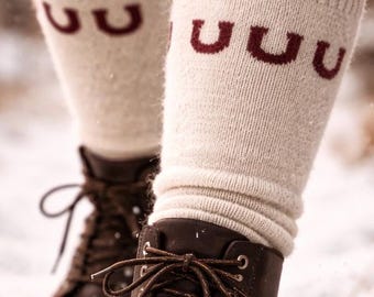 May include: Close-up of brown leather boots with tied laces and cream-colored socks. The socks have a maroon band at the top and maroon "U" shapes. The boots are in a snowy environment.