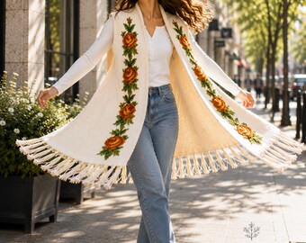 May include: A cream-colored sleeveless knit vest featuring vertical floral embroidery in shades of orange and green along the front panels. The garment has a long, open-front design with a fringed hemline. Text at the bottom reads ANNA FLORIDA.