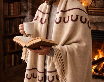 May include: A beige blanket with a burgundy horseshoe pattern and fringe is draped over the shoulders. The blanket is wrapped around a person holding a white mug and an open book. A fireplace and bookshelf are in the background.